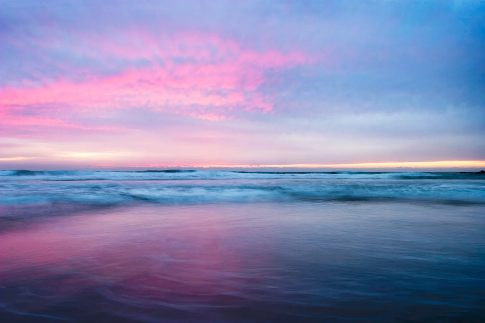 Soft pink and violet sunset over a gentle ocean, with blurred waves rolling to shore.