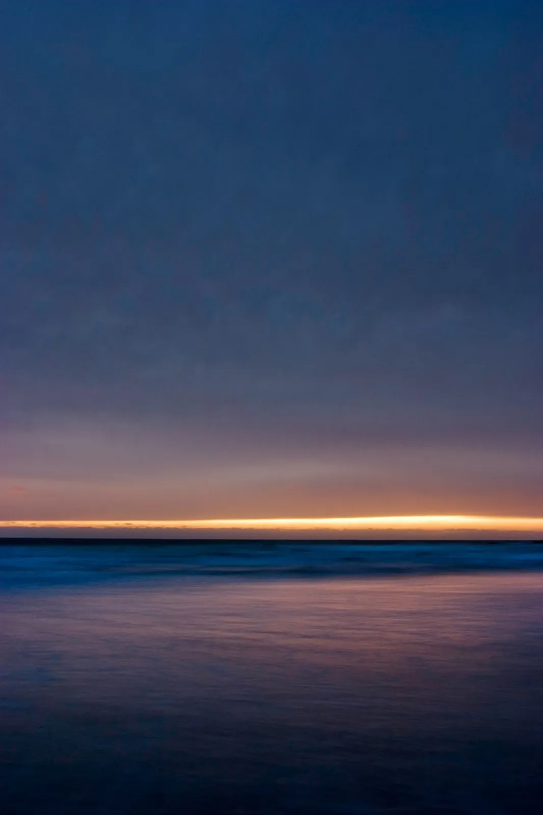 Dusk at the ocean: a thin band of golden horizon light sits between a dark blue cloud-covered sky and a smooth, long-exposure sea.